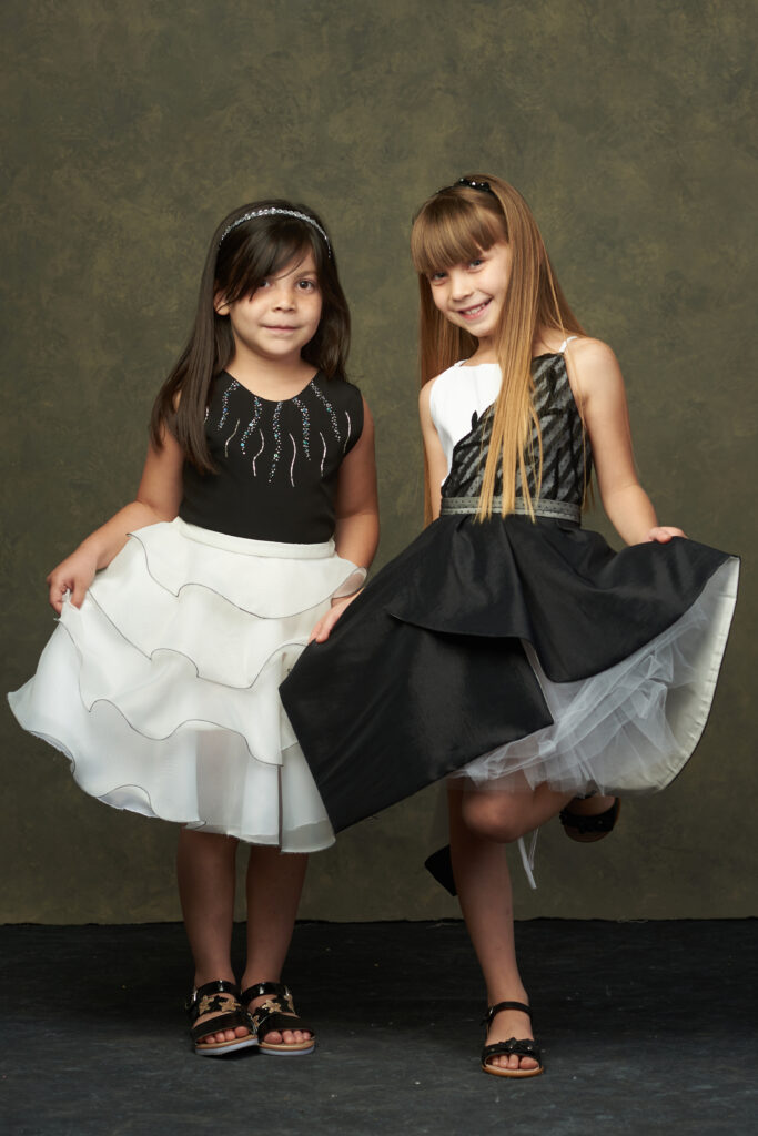 Dos niñas posando en una sesión fotográfica, una lleva una falda negra y blusa blanca, mientras que la otra lleva una falda blanca y blusa negra, sonriendo frente a un fondo gris.