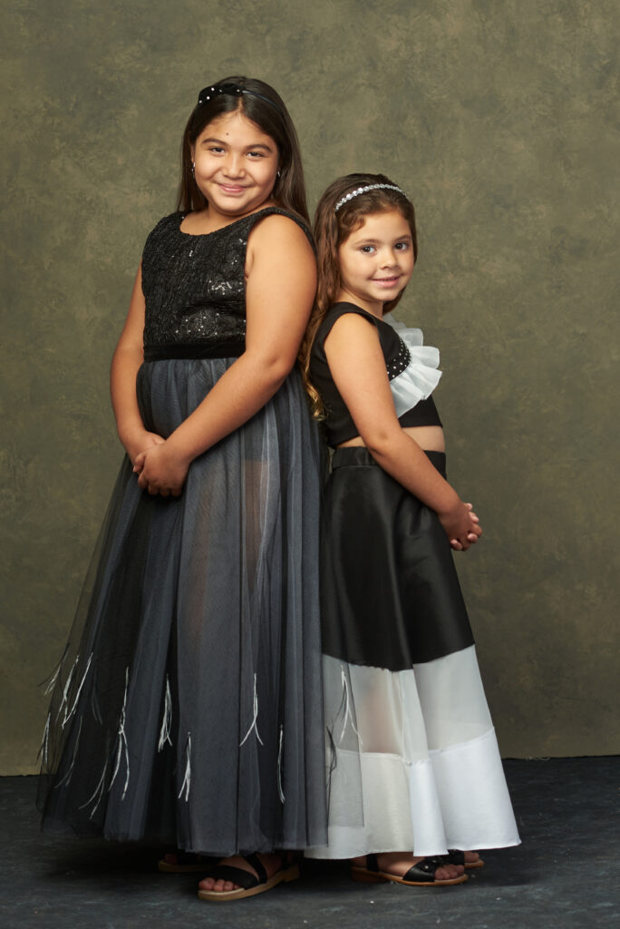 Dos niñas posando sonrientes, una vestida con un vestido negro con detalles brillantes y la otra con un vestido de colores blanco y negro.