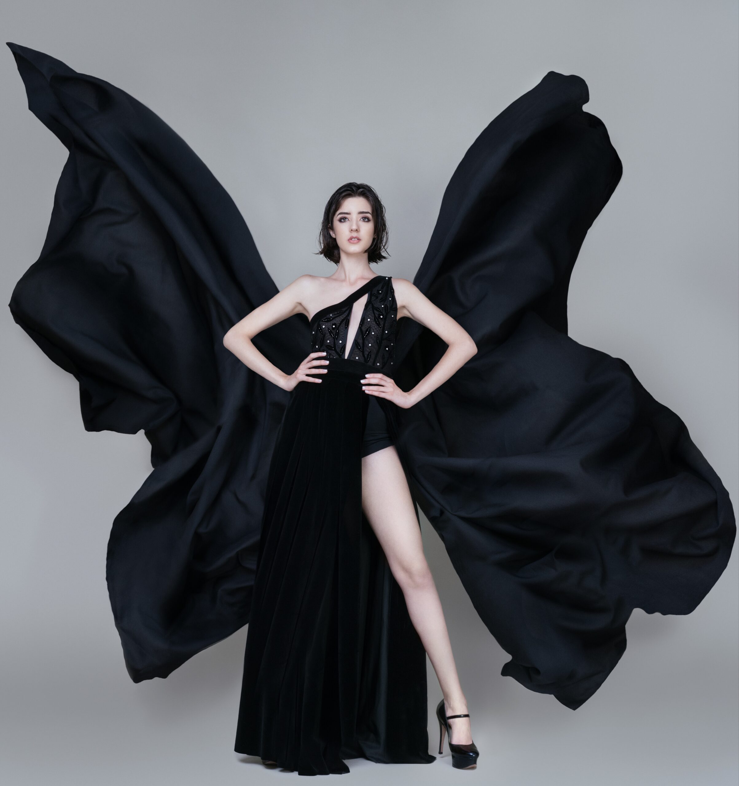 Modelo posando con un vestido negro de alta costura, con mangas que simulan alas y detalles de piedras. Fondo gris sencillo.
