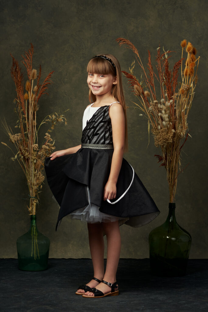 Niña sonriendo con un vestido elegante en tonos negro y blanco, rodeada de flores secas en un fondo texturizado.